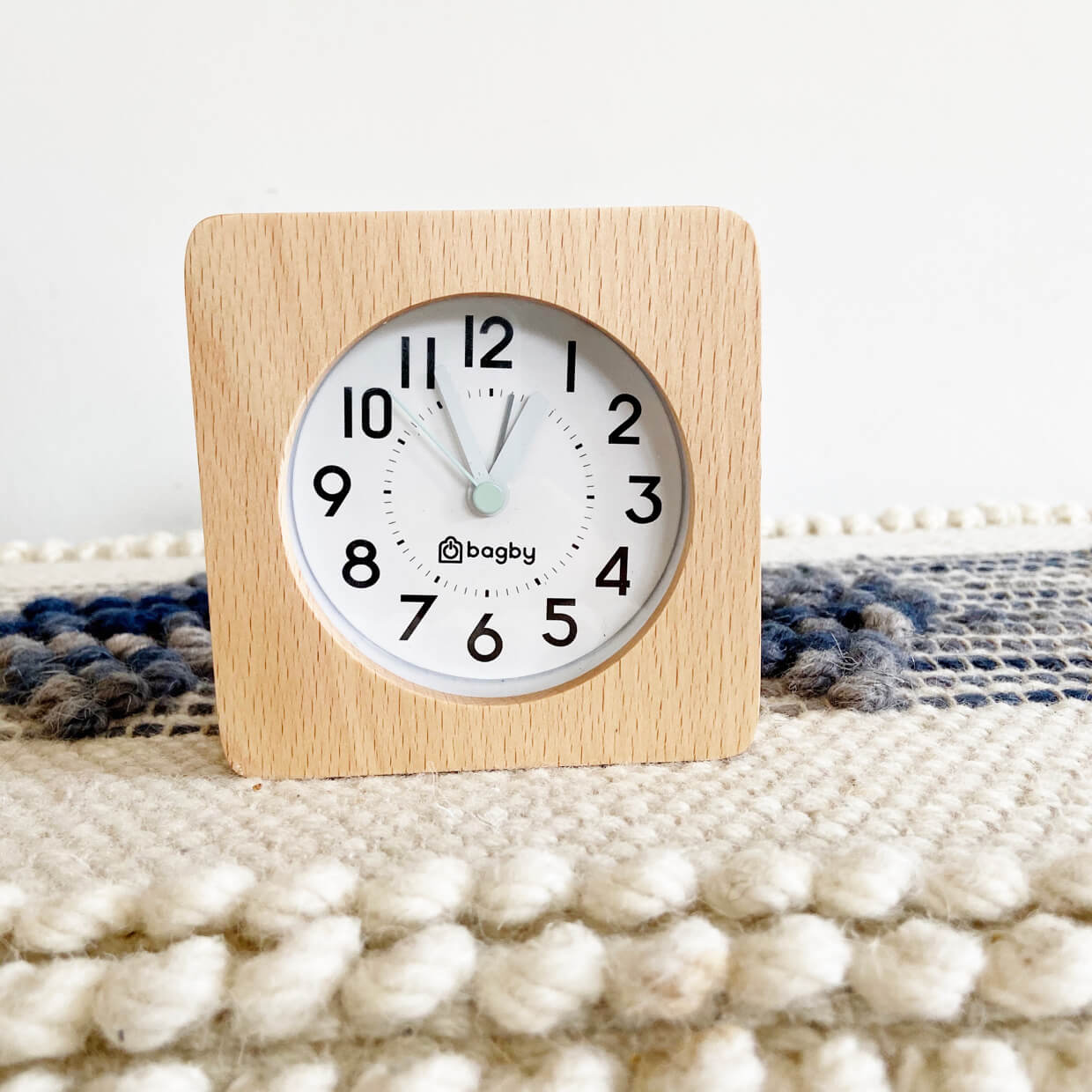 Beech wood Bagby FARMHOUSE alarm clock reading just before one o'clock, set on a woven blue and cream textile surface.