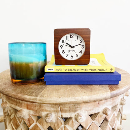 Bagby wooden clock displayed on stacked books and blue box atop carved wooden table beside blue glass with beverage.