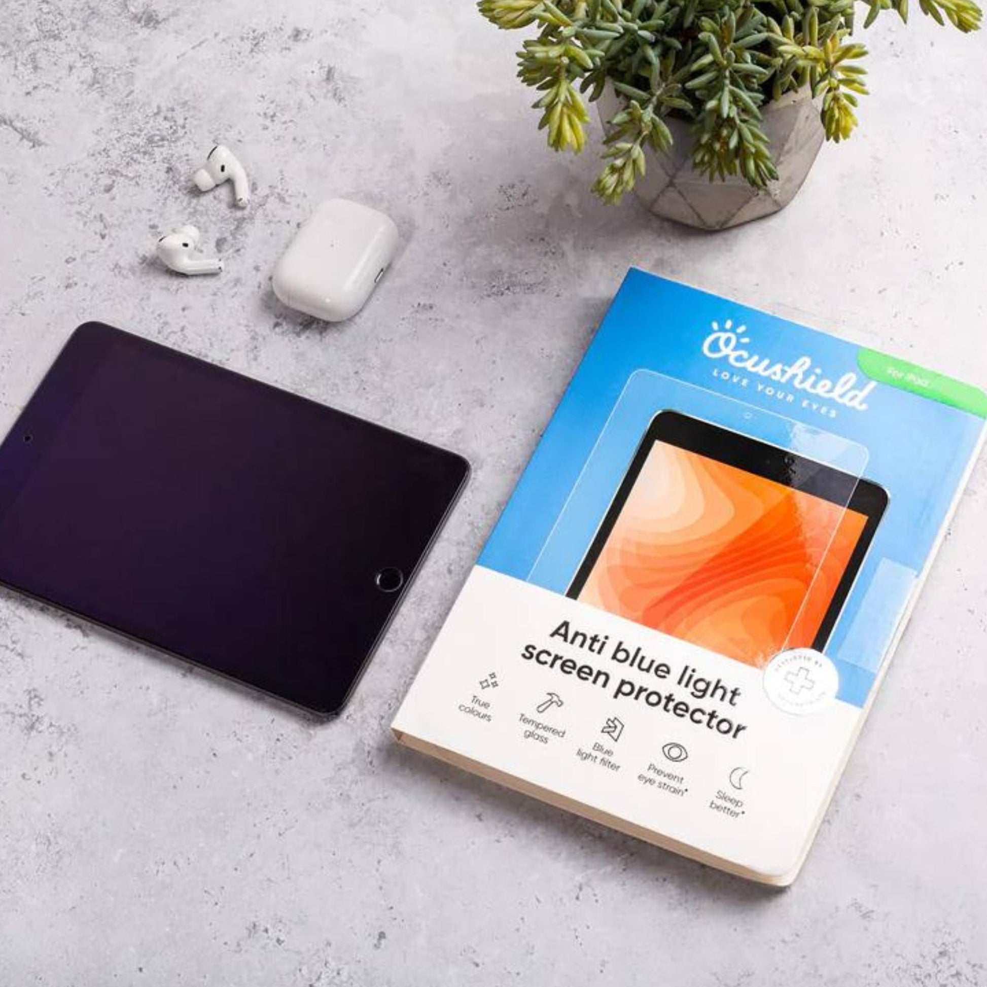 Ocushield blue light screen protector packaging displayed next to tablet, earbuds, and potted plant on gray surface.