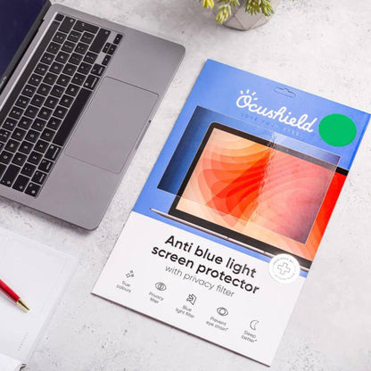 Ocushield blue light screen protector packaging displayed next to open laptop on white desk with potted plant and red pen.