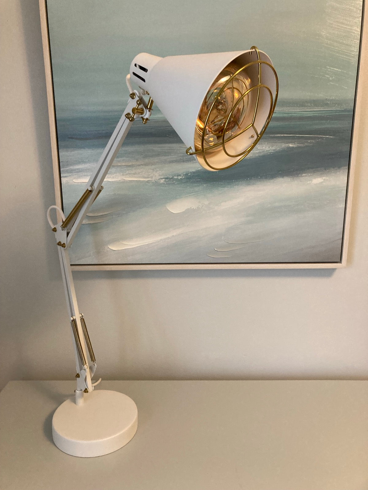 White adjustable-arm desk lamp with a gold wire guard over an amber bulb, standing on a shelf in front of a framed seascape painting.