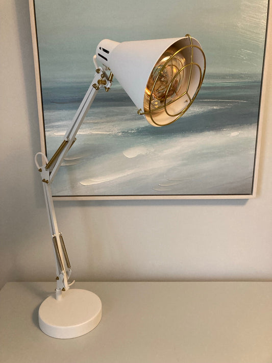 White adjustable-arm desk lamp with a gold wire guard over an amber bulb, standing on a shelf in front of a framed seascape painting.