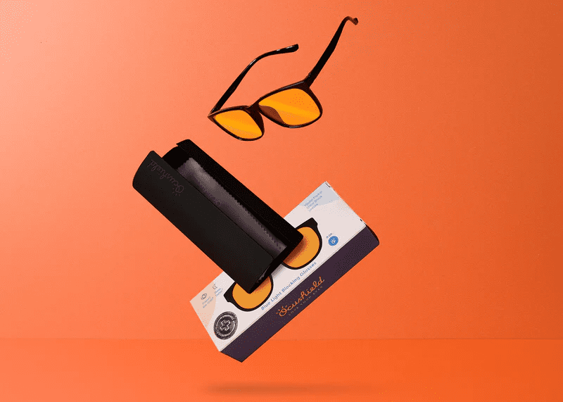 Ocushield Amber blue light glasses with black frames and case displayed against orange background, image 9 of 26.