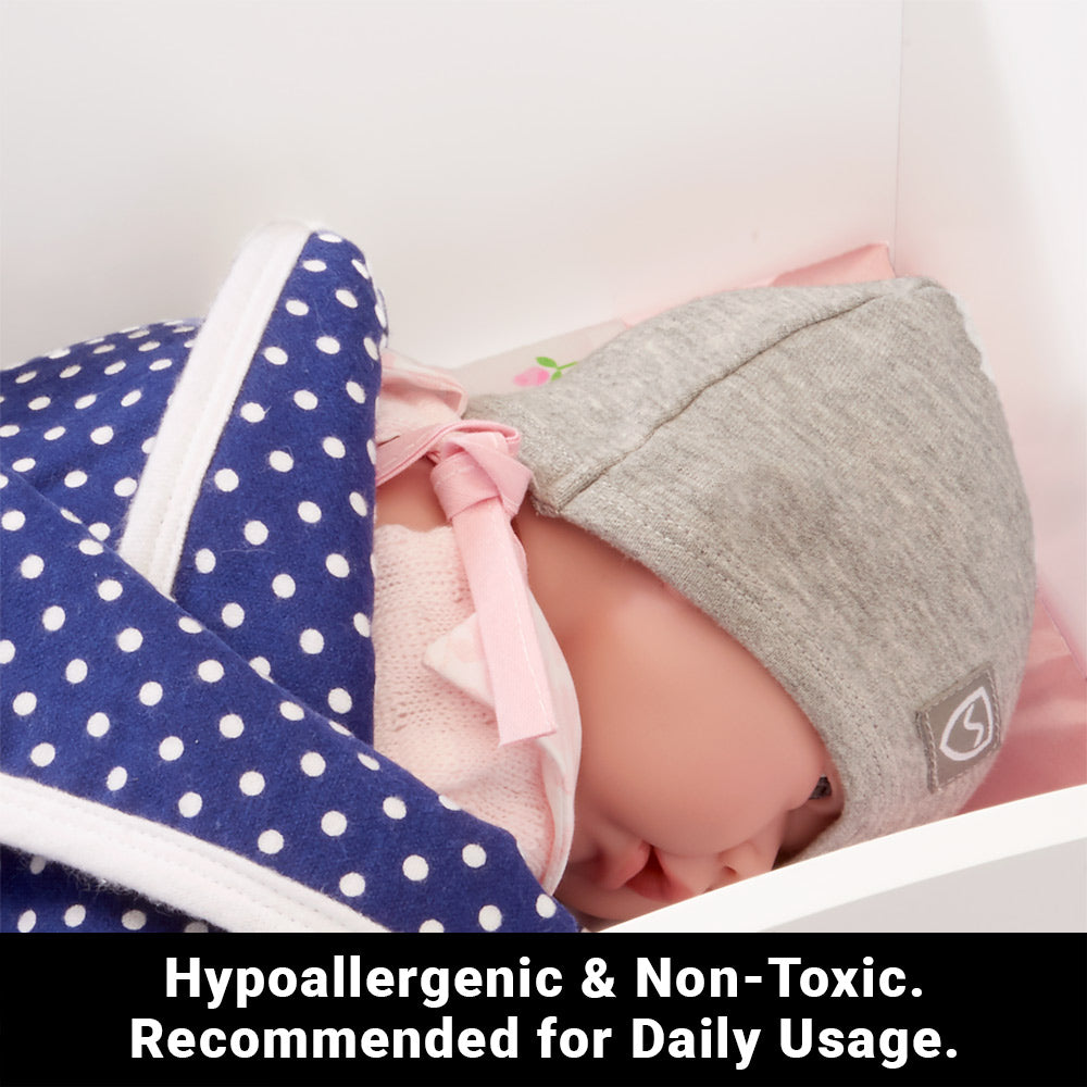 Sleeping baby wearing gray SYB Baby Bundle beanie and navy polka dot blanket with pink ribbon, surrounded by soft pink bedding.