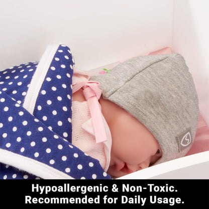 Sleeping baby wearing gray SYB Baby Bundle beanie and navy polka dot blanket with pink ribbon, surrounded by soft pink bedding.