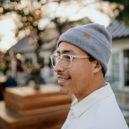 Man wearing gray SafeSleeve Beanie with clear glasses, smiling outdoors with blurred holiday lights background.