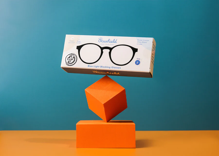 Ocushield Clear All-Day Blue Light Glasses boxed packaging balanced on orange geometric blocks against turquoise background.