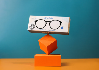 Ocushield Clear All-Day Blue Light Glasses boxed packaging balanced on orange geometric blocks against turquoise background.