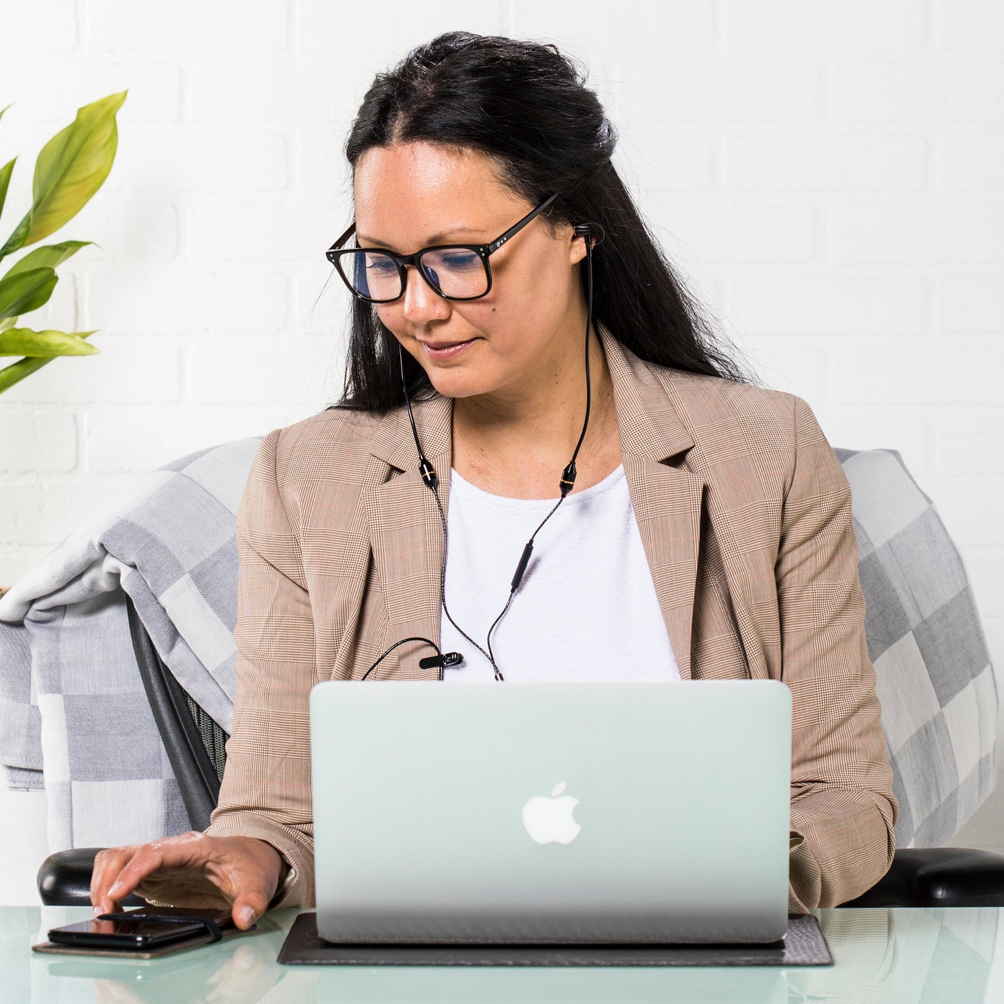 Woman wearing glasses and headset working at laptop on SYB Laptop Pad, seated at desk with plant nearby.