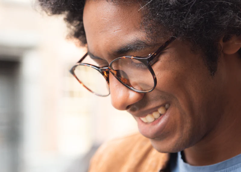 Young man wearing tortoiseshell Ocushield Clear blue light glasses, smiling while looking down outdoors.
