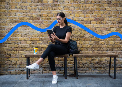 Woman wearing Ocushield Clear blue light glasses while reading tablet on bench against brick wall with blue wavy line mural.