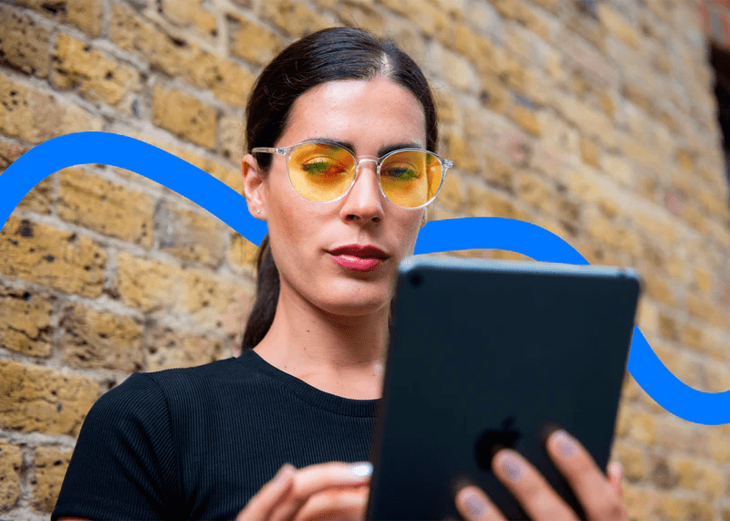 Woman wearing Ocushield Amber blue light glasses with yellow-tinted lenses, holding a tablet device against a brick wall background.