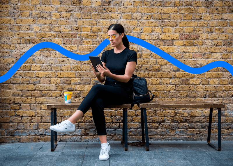 Woman wearing Ocushield Amber blue light glasses sitting on bench against brick wall, holding tablet.