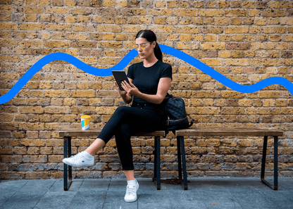 Woman wearing Ocushield Amber blue light glasses sitting on bench against brick wall, holding tablet.