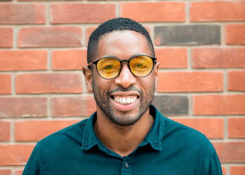 Man wearing Ocushield Amber blue light glasses with yellow-tinted lenses against a brick wall background, smiling at camera.