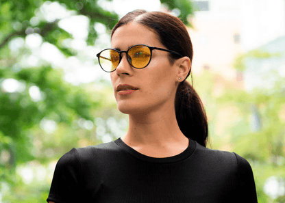Woman wearing Ocushield Amber blue light glasses with amber-tinted lenses and black frames, looking to the side outdoors.