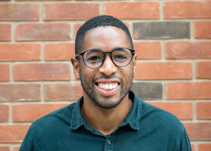 Man wearing Ocushield Clear All-Day Blue Light Glasses with dark frames against a brick wall, smiling at camera.