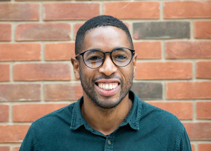 Man wearing Ocushield Clear All-Day Blue Light Glasses with dark frames against a brick wall, smiling at camera.