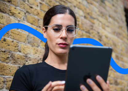 Woman wearing Ocushield Clear All-Day Blue Light Glasses while holding a tablet against a brick wall.