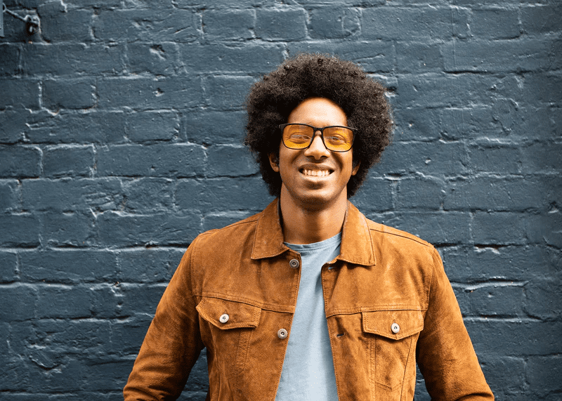Man wearing Ocushield Amber dark blue light glasses and tan jacket smiling against blue brick wall.