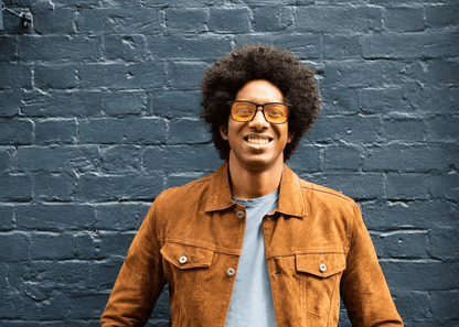 Man wearing Ocushield Amber dark blue light glasses and tan jacket smiling against blue brick wall.