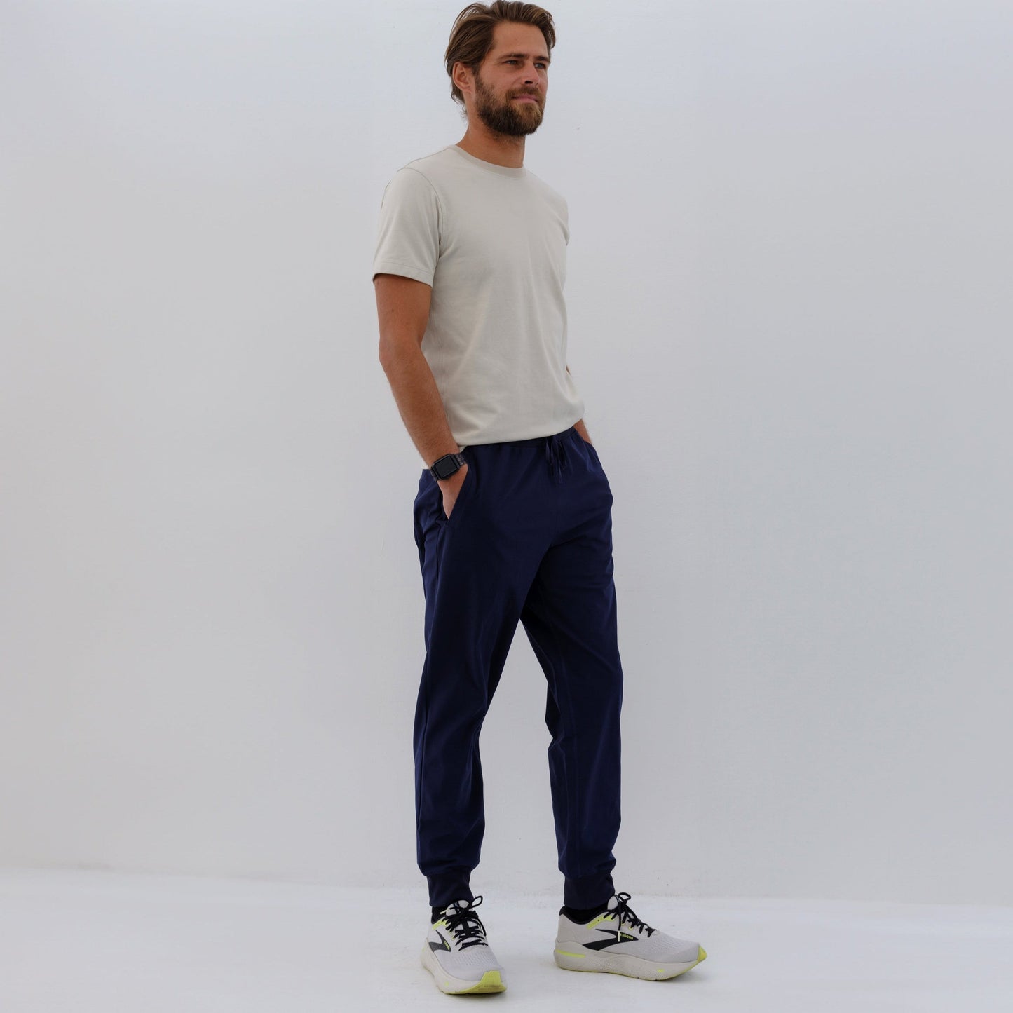 Man wearing beige t-shirt and navy HAVN WaveStopper™ Joggers with white and neon sneakers, standing in side profile against white background.