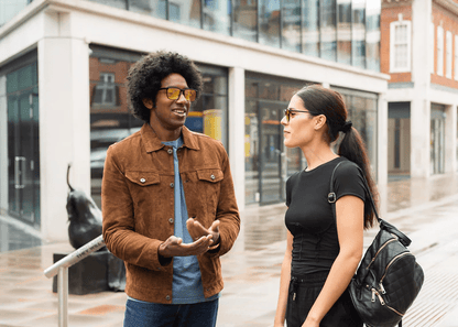Man wearing Ocushield Amber blue light glasses in brown jacket chats with woman on urban street.