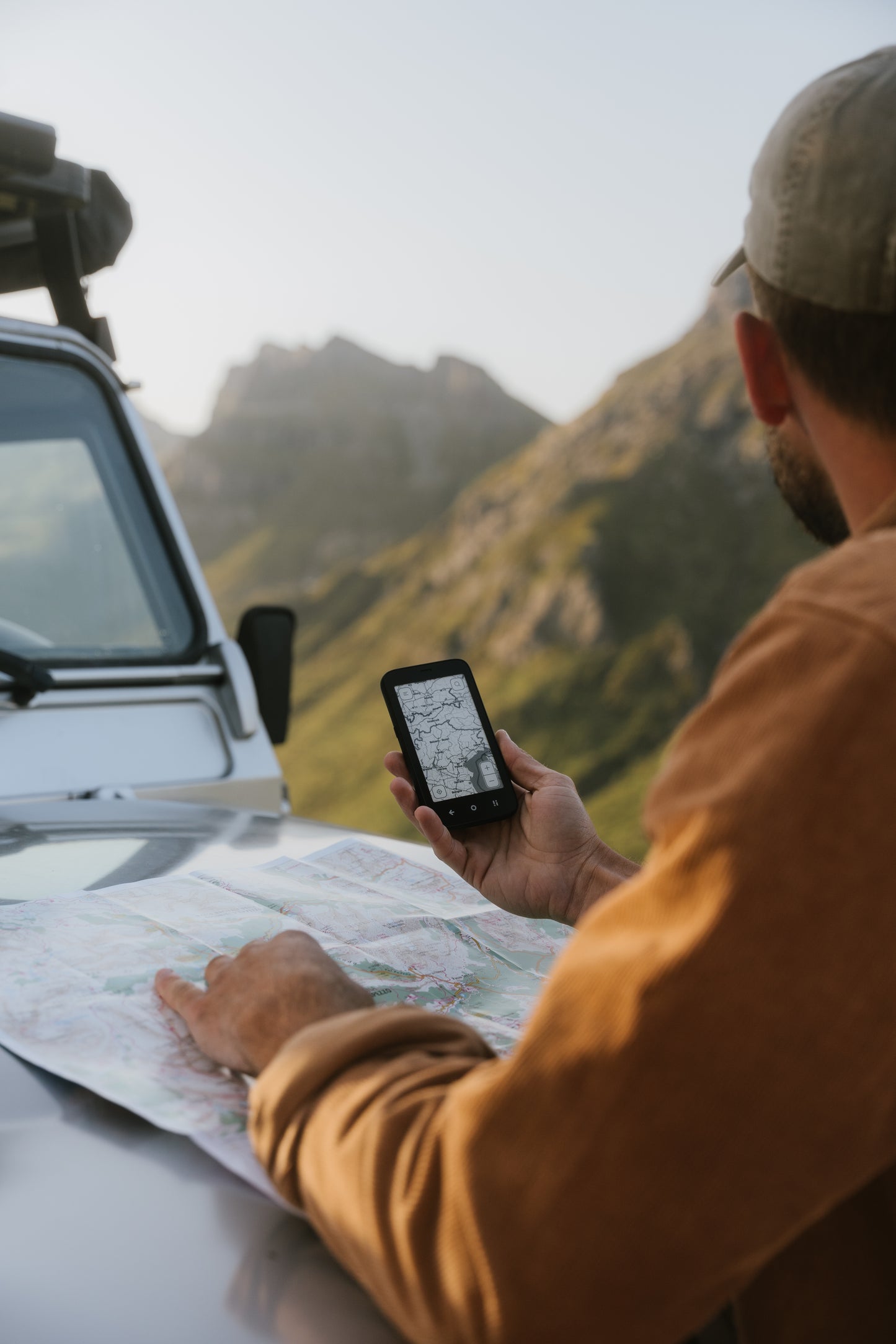 Person holding Mudita Kompakt phone displaying map while viewing paper map near vehicle in mountainous landscape.