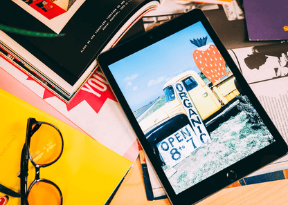 Ocushield Amber blue light glasses displayed on tablet showing beach scene, surrounded by colorful magazines and yellow glasses on desk.