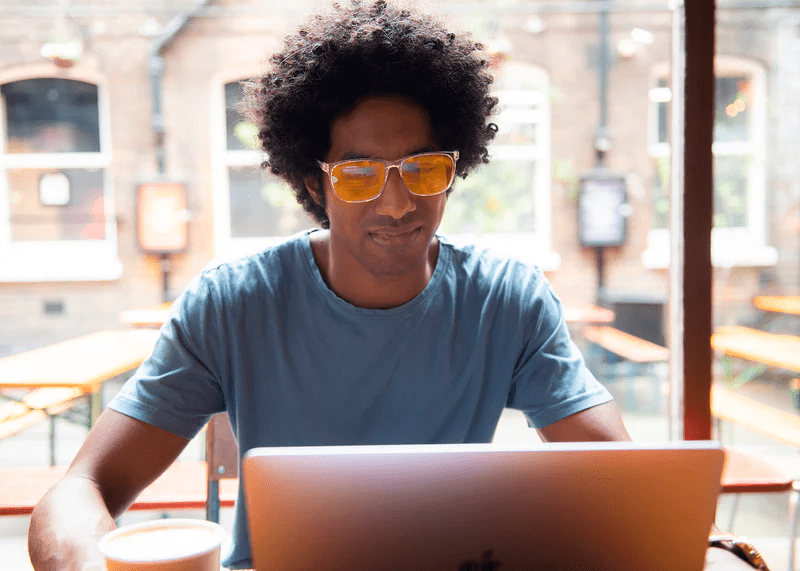 Man wearing Ocushield Amber blue light glasses while using laptop by bright window in café setting.