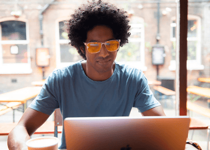 Man wearing Ocushield Amber blue light glasses while using laptop by bright window in café setting.
