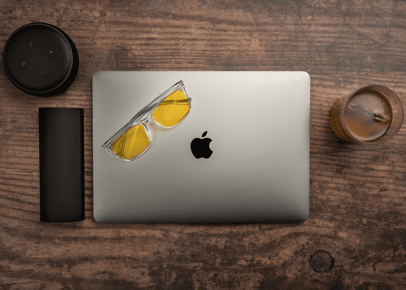 Ocushield Amber blue light glasses with yellow-tinted lenses resting on a MacBook laptop, styled on a wooden desk with coffee and tech accessories.