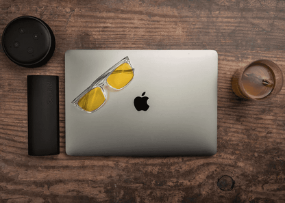 Ocushield Amber blue light glasses with yellow-tinted lenses resting on a MacBook laptop, styled on a wooden desk with coffee and tech accessories.