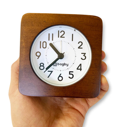 Hand holding a Bagby analog alarm clock with wooden square frame, white face, and dark hands showing 10:10.