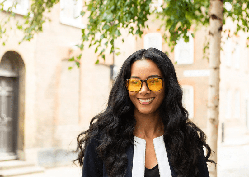 Woman wearing Ocushield Amber blue light glasses with yellow-tinted lenses and black frames, smiling outdoors against a building.