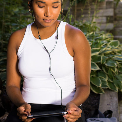 Woman wearing white tank top holding tablet with SYB Tablet Pad, earbuds in, outdoors with plants in background.
