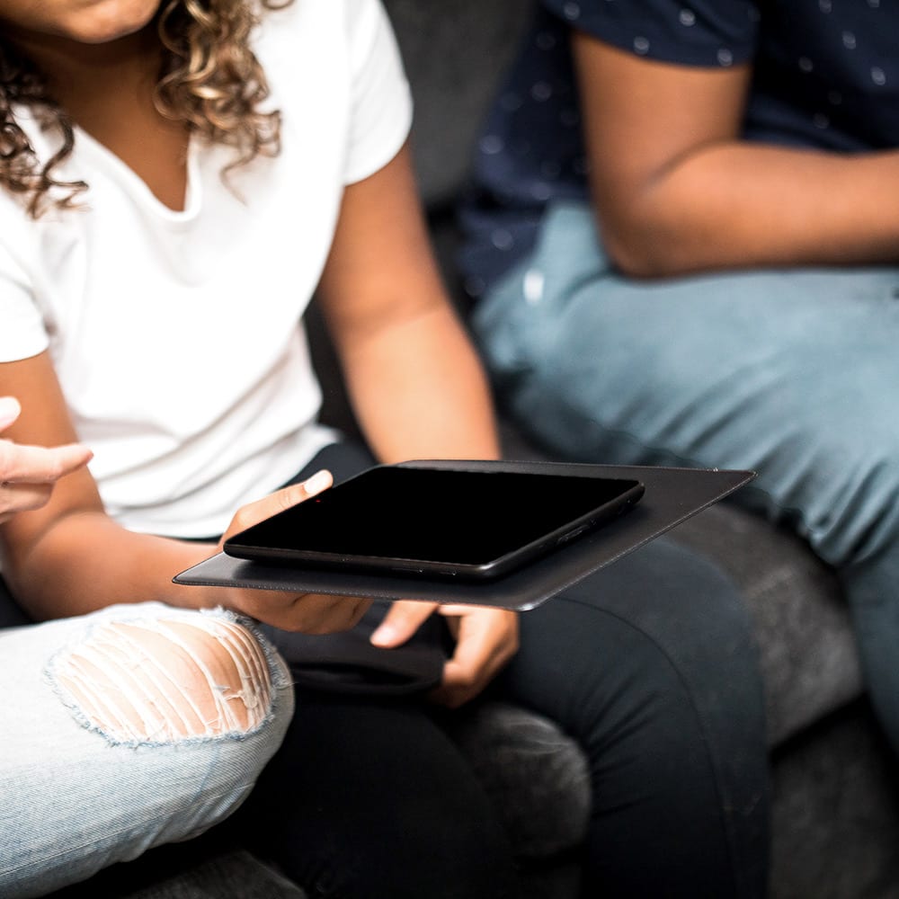 Two people sitting together holding a black SYB Tablet Pad with a smartphone on top, demonstrating the product in use.