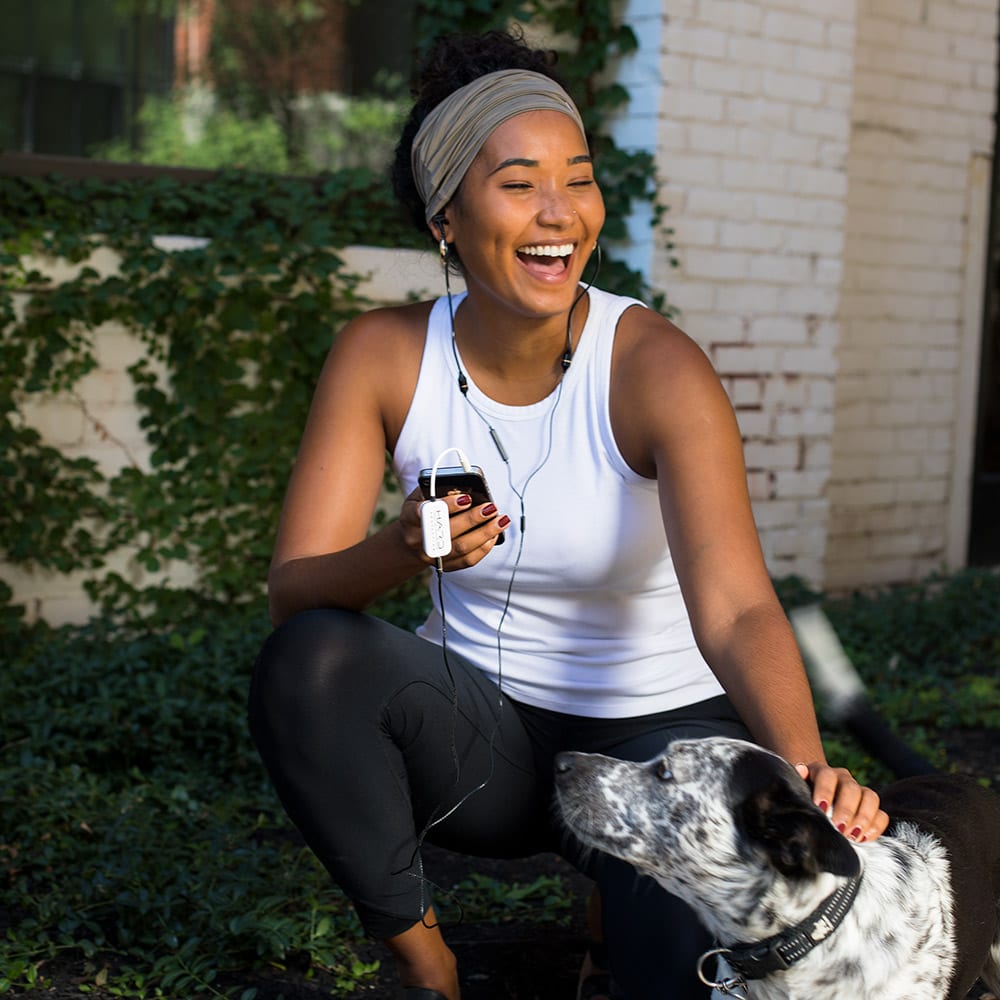 Woman wearing SYB H.A.R.D. headset device while exercising outdoors with dog, smiling.