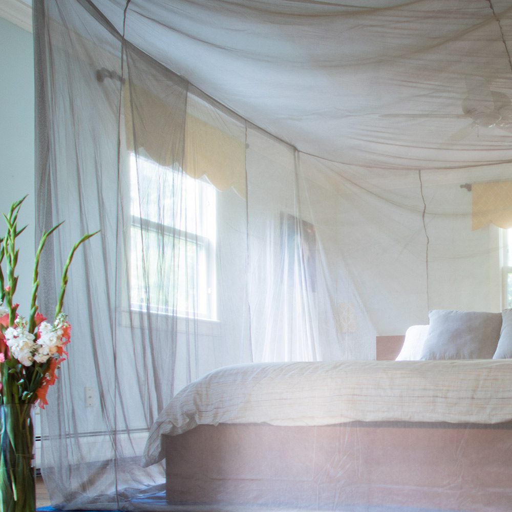 SYB Faraday Bed Canopy draped over bed with sheer mesh fabric filtering sunlight, flowers on bedside table.