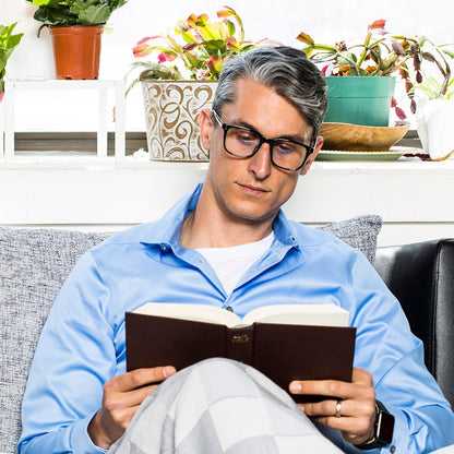 Man wearing SYB Bluelight Glasses and blue shirt reading book on couch with potted plants on shelf behind him.