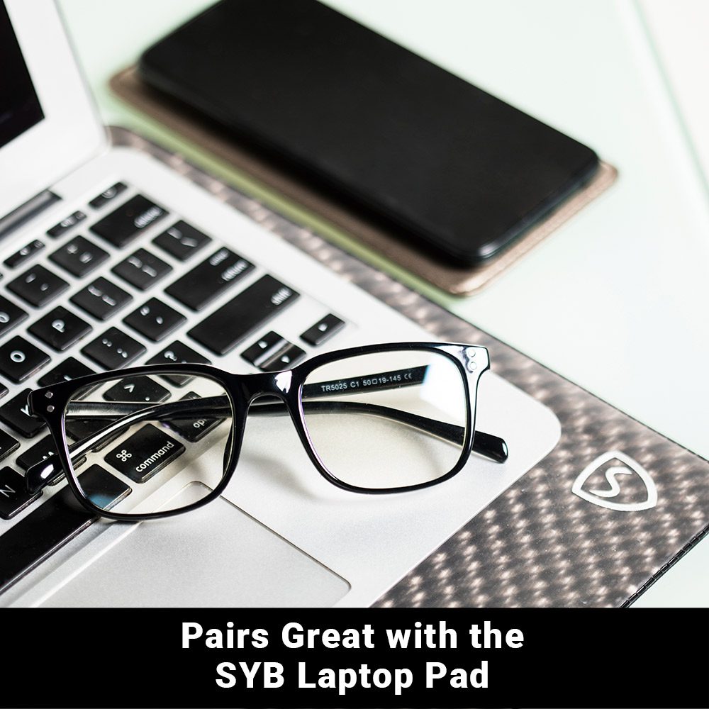 SYB Bluelight Glasses resting on laptop keyboard next to smartphone and mouse pad.