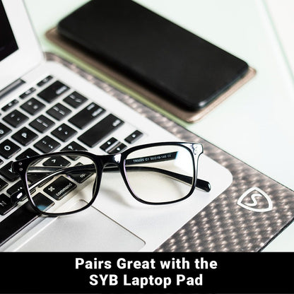 SYB Bluelight Glasses resting on laptop keyboard next to smartphone and mouse pad.