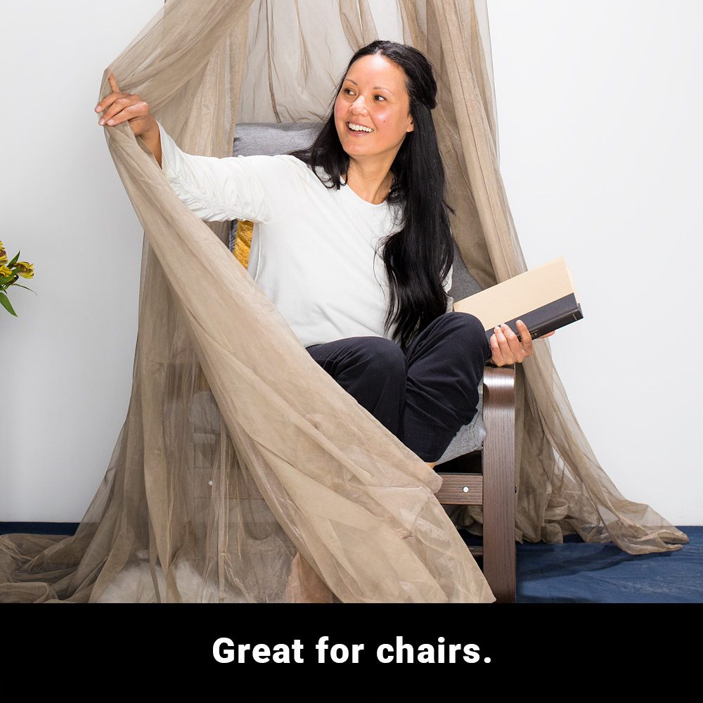 Woman sitting in chair under SYB Faraday Serenity Canopy, holding book, draping sheer fabric with hand, smiling.